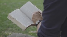 Young man reads the Bible on a bench under a tree outside