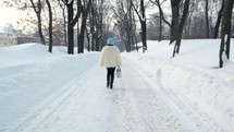 Beautiful woman in white fur coat walking in park. It is snowing. Slow motion. Winter forest. Trendy girl alone.