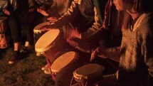 Bunch of friends sit by a fire and play drums in peaceful summer evening, detail of hands drumming on Djembe
