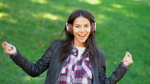 Happy mixed race young woman listening to music on headphones. Hispanic hipster girl dancing to rhythm and singing along melody in the park.