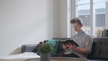 A man sitting on a couch in front of a window reading a Bible 