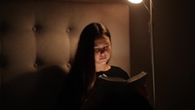 teen girl sitting by a lamp in a dark room reading a Bible 