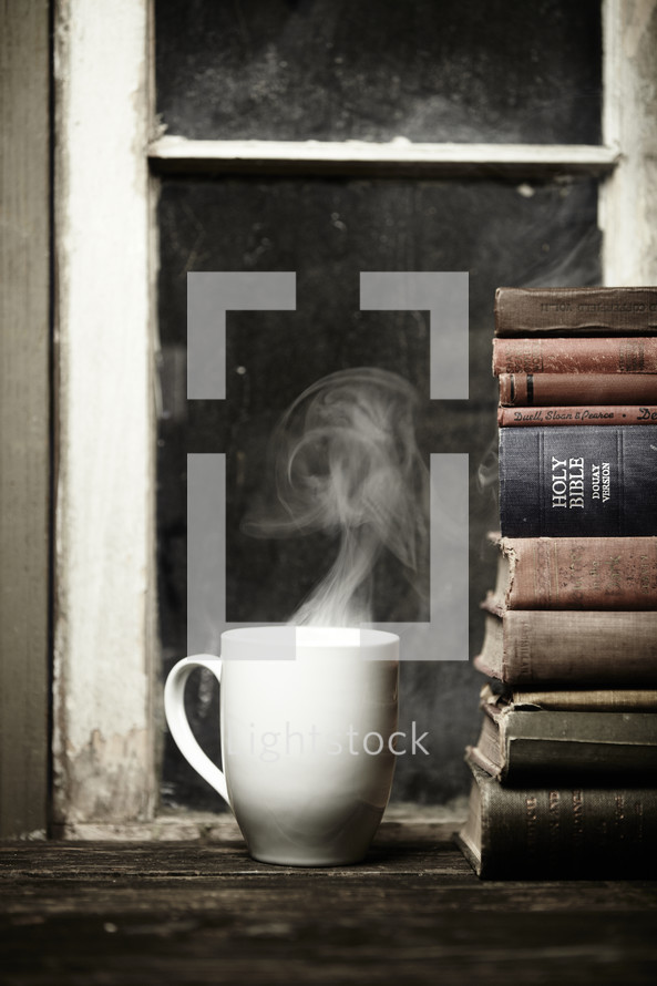 Steaming cup of coffee on a wood table next to a stack of books and bible