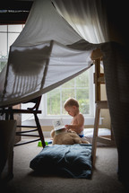 A toddler reading a book under a tent made of sheets.