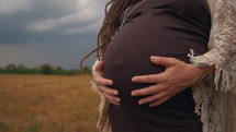 Pregnant woman in a black long dress and dreadlocks against the background of yellow wheat field and stormy sky. Nature, autumn or summer season. Boho style lady touching her big belly. Slow motion