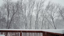 Snowflakes falling down on a wooden deck in the winter time with bare trees in the background.