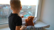 Child dreams concept. Friendship. Lonely little boy hugging teddy bear, looking out window, standing at home alone, happy child waiting for parents. Toddler child sitting at window sill, stay at home.