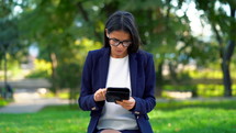 Successful business woman with tablet computer on green park background. Beautiful girl in formal wear and glasses surfing internet or working on electronic device.