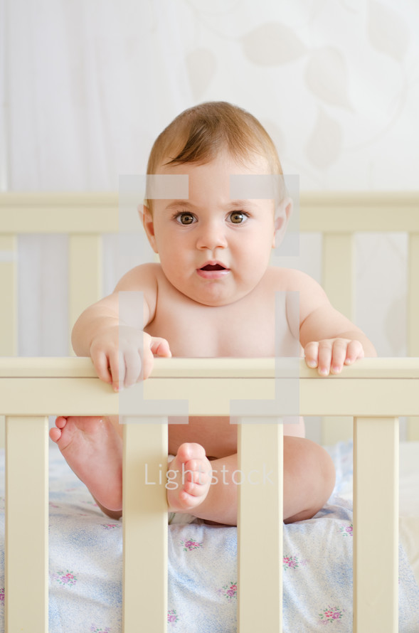 Infant sitting in a crib, holding on to the rail.