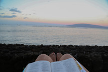Feet of a woman reading a Bible on a beach. 