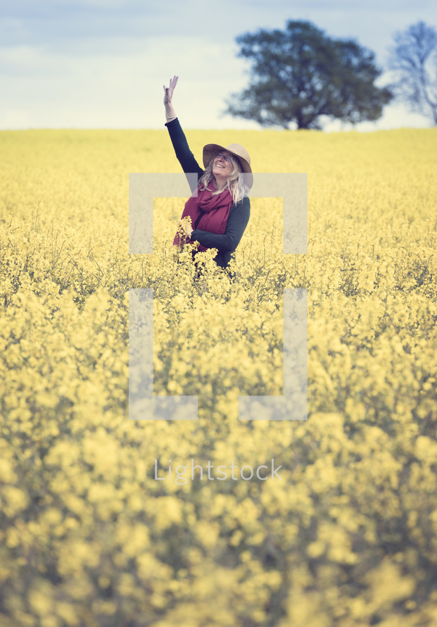 A woman stands in a field of yellow flowers with her hand raised to the sky.