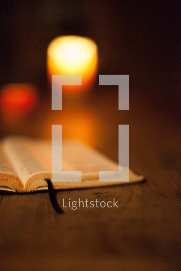Open Bible and lit white votive candle on wooden table.