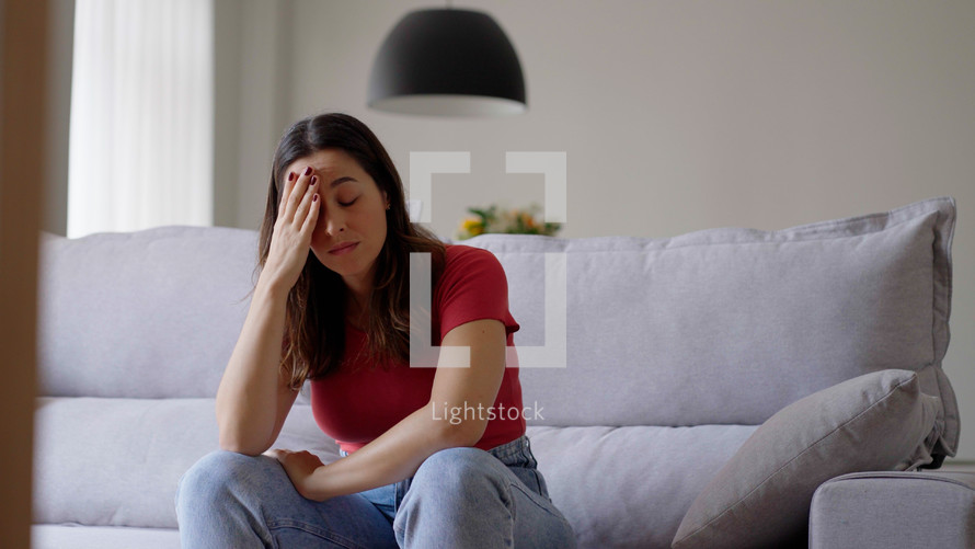 Nervous young woman, feeling unsure about hard decision, sitting alone on sofa. Depressed lady regretting mistake abortion, suffering from psychological problem trouble at home.
