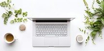 Laptop with cup of coffee and green leaves on white background, top view