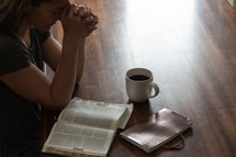 a woman sitting at a table praying and reading a Bible 