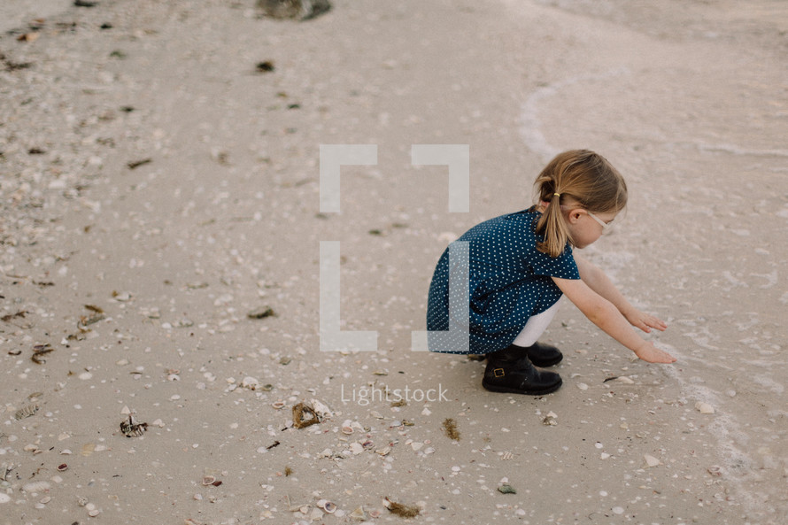 a toddler girl in a dress on a beach reaching for the ocean 