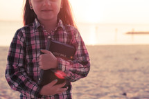 young girl happy, smiling, standing on the beach holding her bible at sunset
