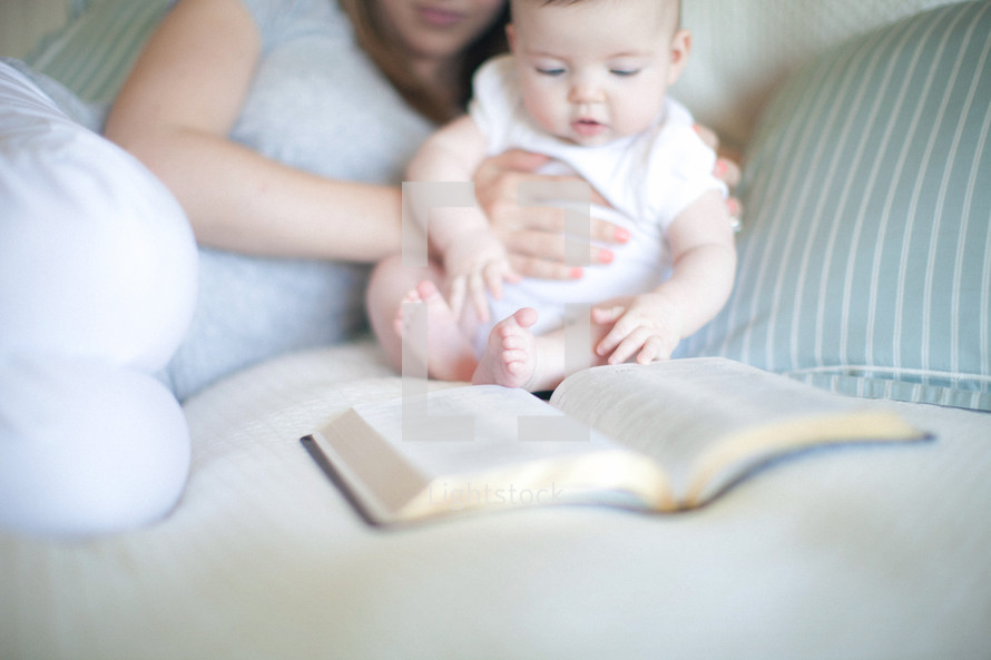 Mother holding infant daughter on the bed looking at an open Bible.