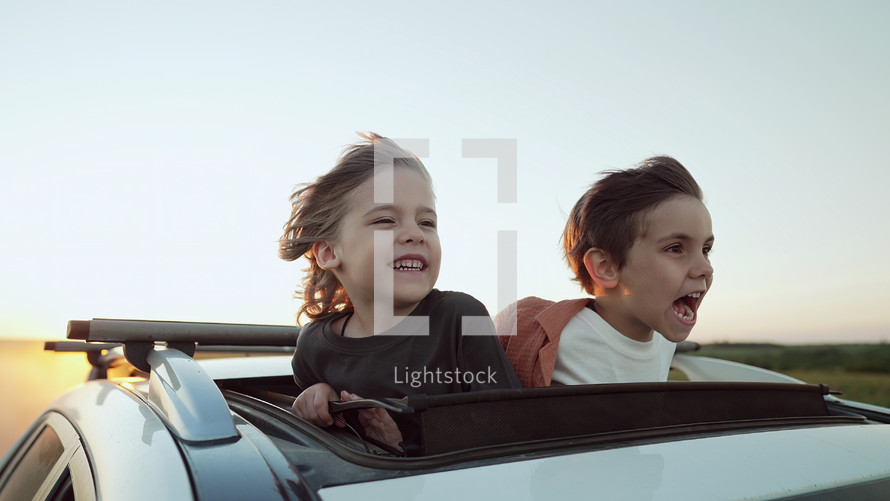 Adorable happy little kids stands in open car sunroof during road trip in countryside, rural landscape at summer. Concept of family leisure, active traveling. High quality