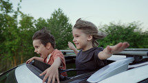 Adorable happy little kids stands in open car sunroof during road trip in countryside, rural landscape at summer. Concept of family leisure, active traveling. High quality