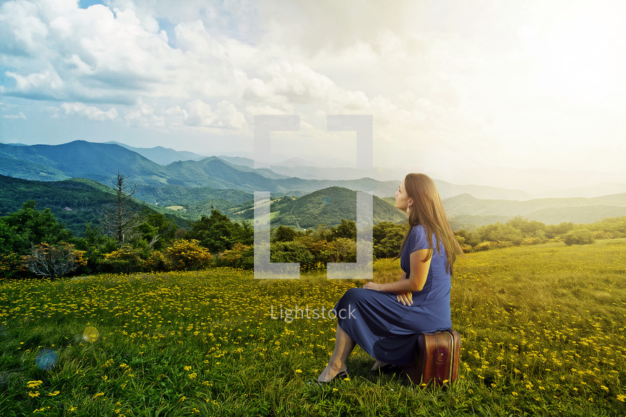 Woman sitting on a suitcase in a grassy meadow with flowers, overlooking a mountain range.