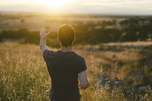 young man with a raised hand standing in a field at sunset 