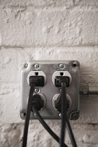 Four electrical cords plugged into an electrical outlet mounted on a brick wall.