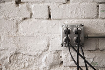Four cords plugged into an electrical outlet mounted on a brick wall.
