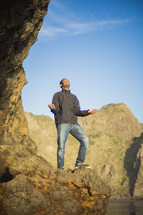 African American man standing on rock cliff with raised hands 