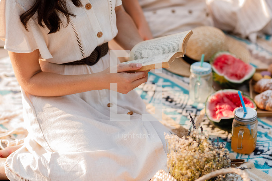 cropped photo of young woman in dress reading book while enjoying picnic in summer park. Sitting on blanket. Croissants, watermelon and cold summer drink.