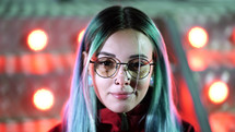 Hipster girl with blue dyed hair, golden sequins as freckles. Woman with nose piercing, transparent glasses, ears tunnels, unusual hairstyle stands in amusement night park