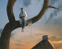 a man reading a Bible sitting on a tree branch next to a rope 