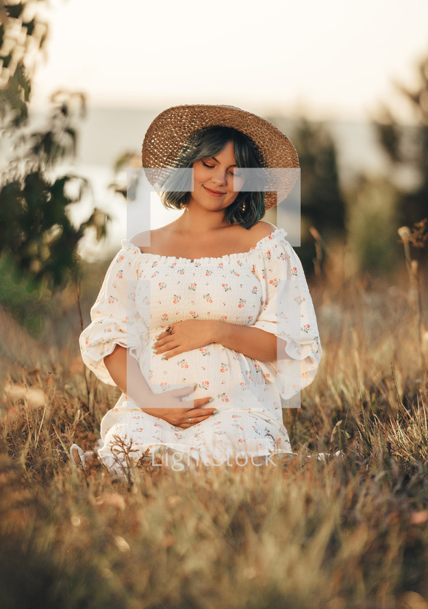 Happy Mother Carrying Child, Expecting For Baby. Amazing Motherhood. Pregnant Woman in beautiful dress and straw hat sitting on grass at nature rural meadow. High quality 