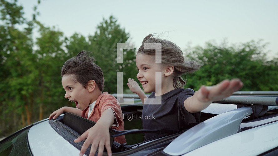 Adorable happy little kids stands in open car sunroof during road trip in countryside, rural landscape at summer. Concept of family leisure, active traveling. High quality