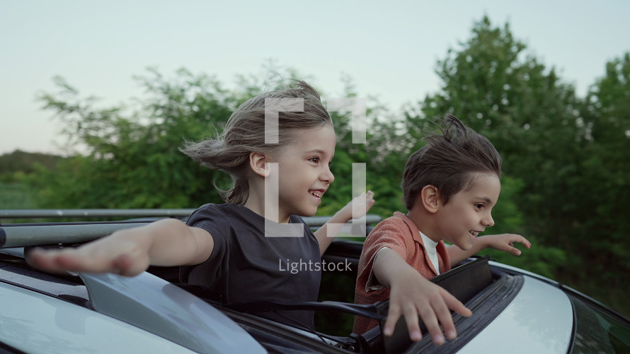 Adorable happy little kids stands in open car sunroof during road trip in countryside, rural landscape at summer. Concept of family leisure, active traveling. High quality