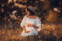 Happy Mother Carrying Child, Expecting For Baby. Amazing Motherhood. Pregnant Woman in beautiful dress and straw hat sitting on grass at nature rural meadow. High quality 