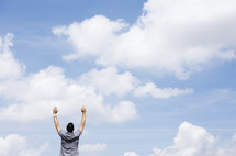 man with raised arms standing outdoors with blue skies as background 