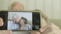 Senior caucasian couple holding up a smartphone to the camera with an image of themselves kissing then revealing themselves behind themes of love romance technology
