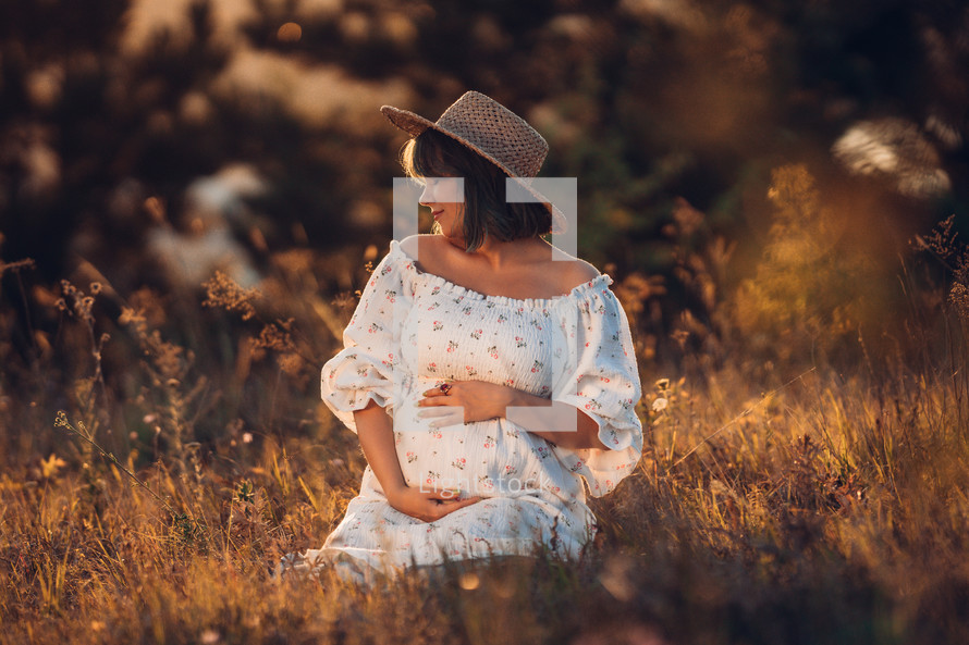 Happy Mother Carrying Child, Expecting For Baby. Amazing Motherhood. Pregnant Woman in beautiful dress and straw hat sitting on grass at nature rural meadow. High quality 