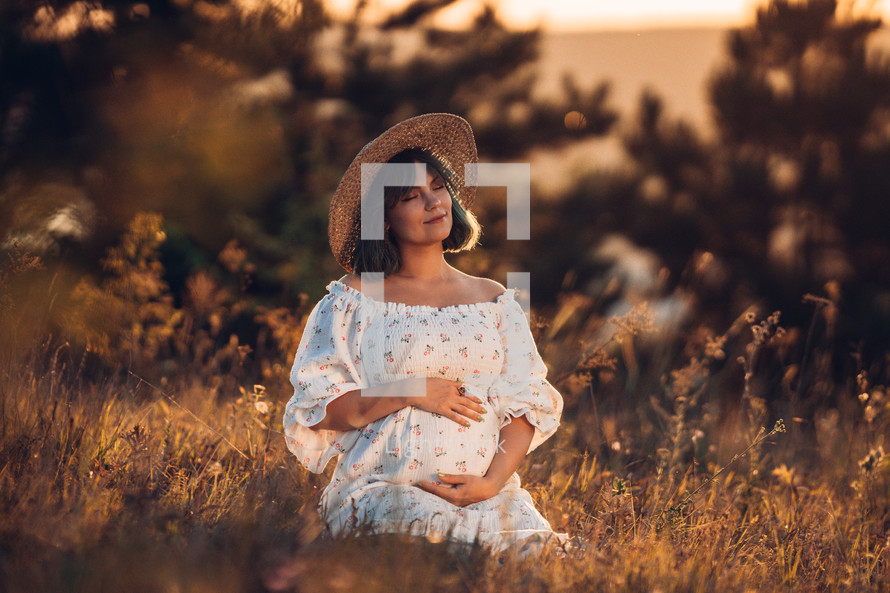 Happy Mother Carrying Child, Expecting For Baby. Amazing Motherhood. Pregnant Woman in beautiful dress and straw hat sitting on grass at nature rural meadow. High quality 