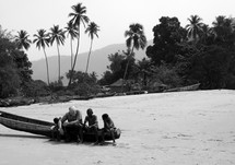 A man and children sit on a canoe on a beach in a tropical setting.