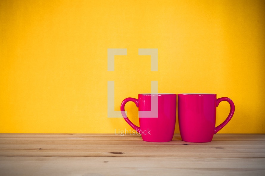 two red mugs on a wood table against a yellow wall 