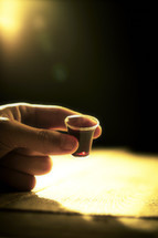 A hand holds a communion cup filled with wine as a light shines bright in the background.