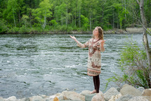woman standing at the shore of a river with her hands raised in praise and worship to God