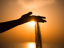 Silhouette of a young girl's hand pouring sand from a beach against a yellow sky at sunset