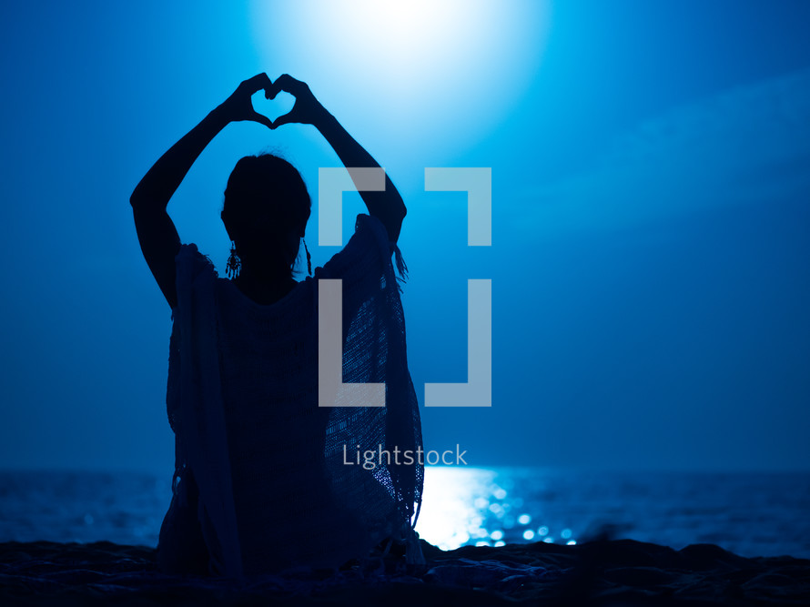 Young girl sitting at night on beach against moon road on sea and made hand heart frame shape silhouette
