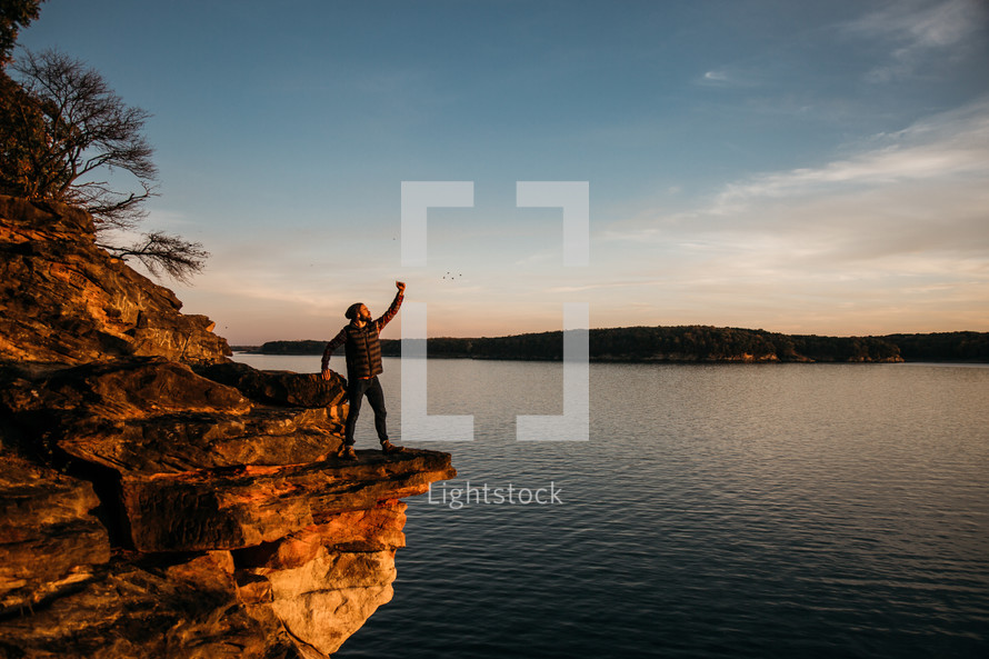 a man with a raised arm standing on the edge of a cliff at sunset 