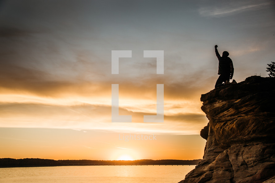 silhouette of a person with a raised fist kneeling at the edge of a cliff at sunset 