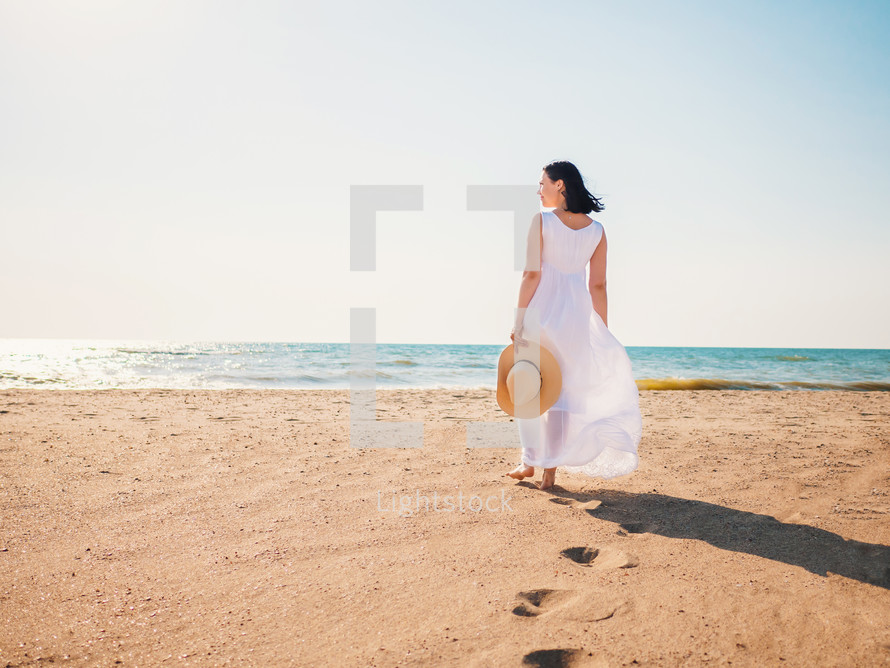 Girl wearing white maxi long dress walking barefoot on the sea shore with hat in hands. Bohemian clothing style.
