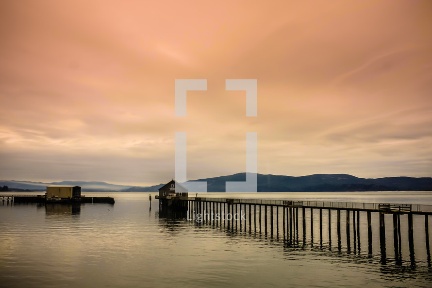 Pier leading to a boathouse in the water with mountains on the horizon at dusk.